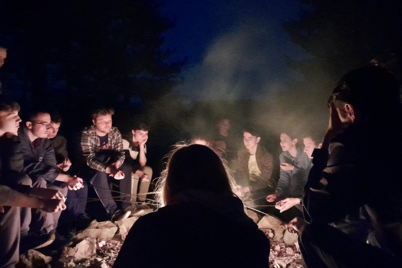 Skupina mladých lidí v noci opéká špekáčky u ohně.